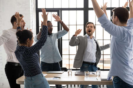 Happy excited millennial employees shouting for joy, raising hands for emotional high fives. Business team of overjoyed professionals cheering, celebrating good sales result, financial goal achievingの写真素材