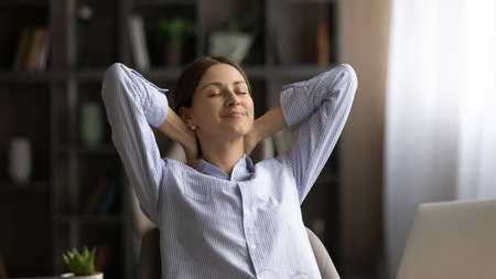 Putting workday on pause. Peaceful young woman relax by desk take break in work meditate with closed eyes. Serene millennial female enjoy moment of rest stretch hands over head on comfy office chairの写真素材