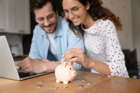 Happy millennial family couple putting coins in piggybank, planning vacation or investments together, saving money for life insurance, managing future expenditures together using computer apps.の写真素材