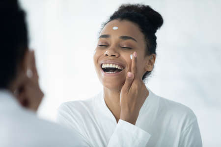 Happy Afro American woman in bathrobe excited with home spa activity, enjoying beauty care procedures, applying facial cream, collagen rejuvenating mask on face at mirror, laughing with closed eyesの写真素材