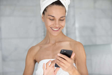 Happy joyful young woman reading text on mobile phone screen after morning shower, using app on cellphone at home, checking messages on social media, chatting online, flirting on dating websiteの写真素材