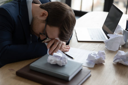 Exhausted office worker resting head on work desk with laptop and crumpled documents, paper drafts....