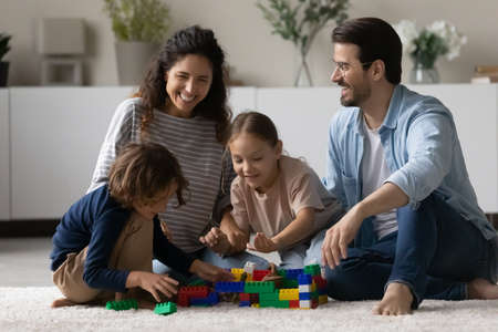 Overjoyed multiethnic parents sit on floor play toys with little biracial kids together. Happy multiracial young family with small ethnic children have fun engaged in playful game activity at home.の写真素材