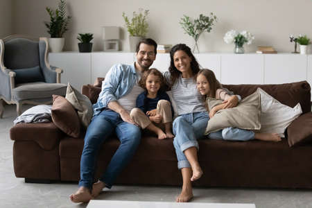 Portrait of smiling young multiracial family with two small kids sit relax on couch in living room. Happy multiethnic parents renters with little children rest at home together. Rental concept.の写真素材