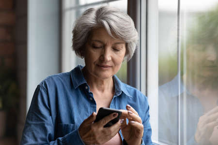 Serious mature woman standing near window with smartphone, share text messages, browse internet...