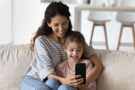 Overjoyed young Latino mother and daughter use cellphone look at screen laugh watch funny video...