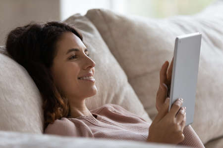 Smiling Latino woman relax on sofa at home look at tablet screen talk speak on video call on gadget....