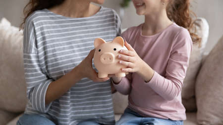 Crop close up of smiling Hispanic mom and biracial daughter hold piggy bank recommend family savings. Happy Latin ethnic mother and teen girl child use piggybank care of budget for money investment.の写真素材