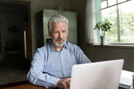 Serious mature Caucasian man sit at desk at home office look at laptop screen work online on gadget....