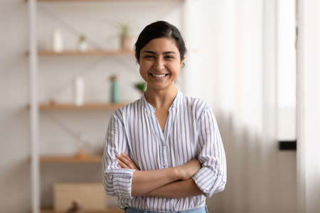 Portrait of happy young Indian female renter tenant stand arms crossed pose in living room at home. Smiling millennial successful confident mixed race ethnicity woman show leadership at workplace.の写真素材