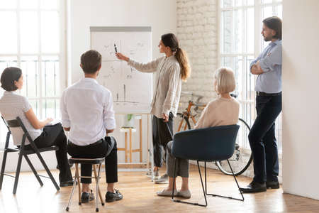 Confident businesswoman coach making flip chart presentation to diverse employees at workshop,...