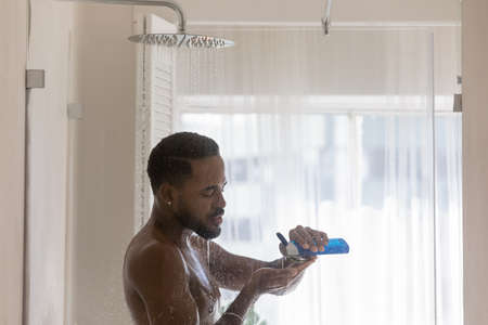 Relaxed attractive young African American guy taking shower, standing under showerhead flowing water, squeezing shampoo, gel, soap from bottle. Body care, haircare, morning hygiene, bath conceptの写真素材