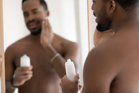 Handsome muscular mixed race Black young man applying aftershave lotion, balm on soft stubble, stylish beard at mirror, holding plastic bottle, touching face. Male beauty care procedures conceptの写真素材