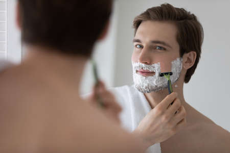 Positive attractive young guy with gel foam on lower face shaving with razor at mirror. Handsome man enjoying morning bath routine, caring for appearance, healthy facial skin. Hygiene, skincare conceptの写真素材