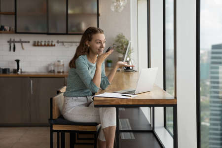 Happy young woman recording audio message on cellphone or holding distant loudspeaker conversation,...
