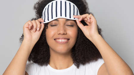 Close up of happy young African American woman isolated on grey studio background wear eye mask go to sleep. Smiling millennial mixed race female get ready for nap or daydream. Relaxation concept.の写真素材