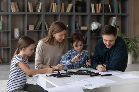 Happy young Caucasian family with two teen children have fun drawing painting together in album. Smiling loving parents enjoy weekend with small teenage kids involved in studying hobby activity.の写真素材