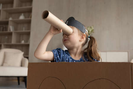 Cute adorable little girl in black pirate hat looking in spyglass, sitting in toy cardboard ship, preschool child kid wearing homemade costume holding cardboard tube as telescope, playing funny gameの写真素材
