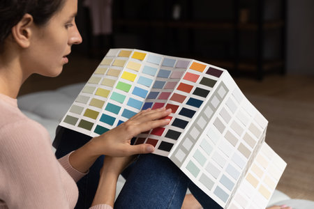 Back rear side view concentrated creative young woman sitting on sofa, holding paint swatches palette, choosing favorite colors for renovating apartment, planning repairment or interior design.の写真素材
