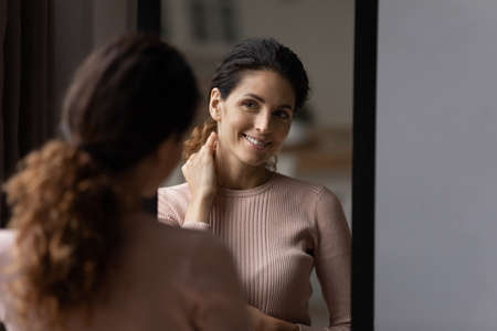 Rear back smiling 30s hispanic caucasian woman looking at mirror, checking appearance before leaving apartment, getting ready for job or walk, satisfied with neat fit look, admiring herself at home.の写真素材