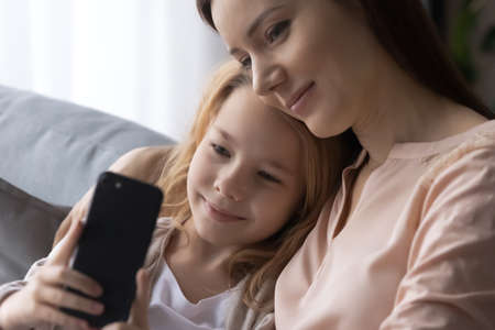 Close up of little daughter and smiling adult mommy cuddle on couch look on girl mobile phone screen watch school photo share news with friends at social networks. Mother and preteen child using cellの写真素材