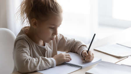 Little pupil. Focused preteen schoolgirl sit at desk in light classroom write exercise in copybook....