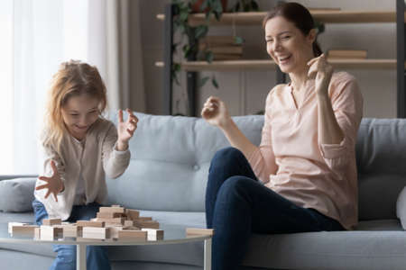 I win. Joyful millennial female foster mother having fun play board game with adopted daughter enjoy funny moment. Happy nanny and laughing preteen girl compete in building tumbling tower at playtimeの写真素材