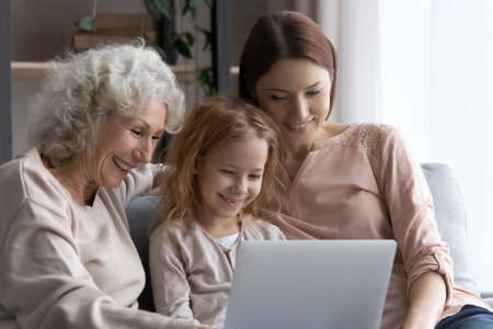 Three age family. Happy little girl daughter grandkid sit on sofa between elderly grandma and adult...
