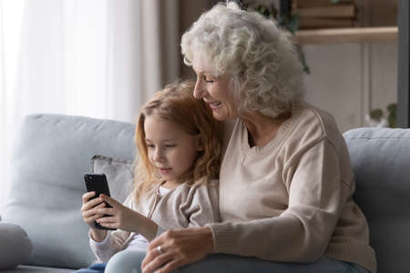 Using cell with grandma. Preteen girl grandkid cuddle on comfy sofa with older grandmother show granny funny photo on phone. Aged hoary grandparent enjoy online mobile game with beloved grandchildの写真素材