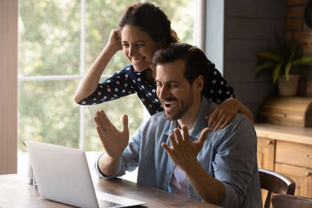 Overjoyed millennial couple using laptop, looking at screen, shouting for joy and laughing. Happy husband and wife getting good surprising news, income, loan or mortgage approval, winning prizeの写真素材