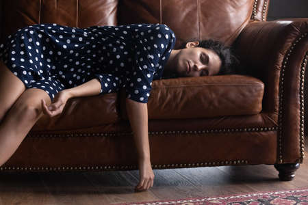 Exhausted Hispanic woman resting on comfortable leather couch at home, sleeping peacefully with hanging hands and legs, feeling fatigue, burnout, suffering from jetlag drowse. Sleep disorder conceptの写真素材