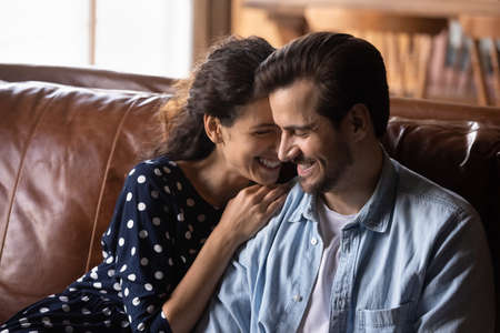 Happy millennial couple enjoying being together in new house, apartment, excited about good news, talking, hugging, laughing, smiling while sitting on luxury leather couch. Love, relationship conceptの写真素材