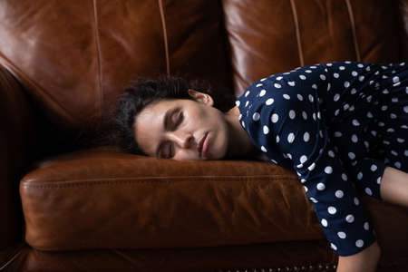 Tired peaceful millennial Latin woman with closed eyes lying on soft cozy sofa, sleeping at daytime,...