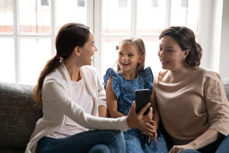 Excited three generations of Caucasian women sit on sofa at home have pleasant webcam virtual event...