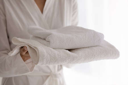Close up shot of young woman in bathrobe going to take shower holding stack of white soft fluffy terry towels. Female client of spa or hotel carry two piece cotton towel set prepare for bath procedureの写真素材