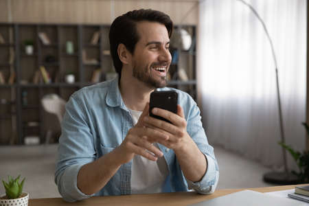 Happy young handsome man holding smartphone in hands, looking in distance, feeling excited of getting amazing news, daydreaming visualizing future, thinking of new challenges or opportunities.の写真素材