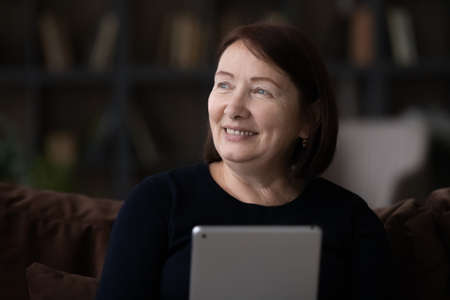 Happy mature 60s woman using tablet at home, sitting on couch, holding digital device, getting good...