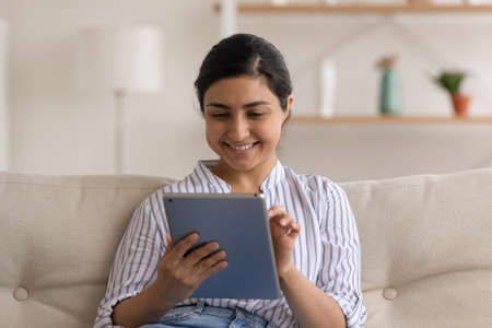 Close up smiling Indian woman using tablet, sitting on couch at home, happy positive young female...