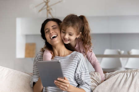 Overjoyed small teen biracial girl child and young Hispanic mother have fun using modern tablet gadget at home. Smiling Latino mom and little daughter speak talk on video webcam call on pad.の写真素材