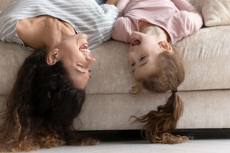 Excited young Hispanic mother and teen daughter lying on couch upside down have fun relaxing at home together. Smiling Latin mom and little girl child sing listen to music enjoy family weekend.の写真素材