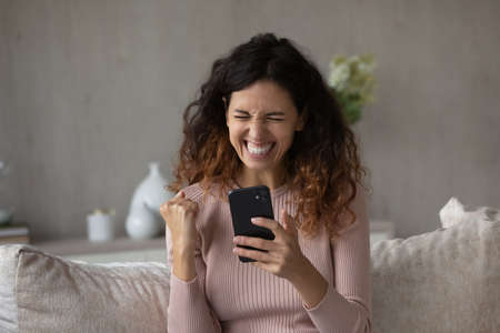 Overjoyed millennial Hispanic woman feel euphoric win online lottery on cellphone. Smiling young...