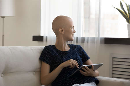 Dreamy positive young hairless woman using digital computer tablet, enjoying playing online games, web surfing information about fighting cancer disease or reading supportive motivational articles.の写真素材