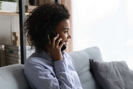 Smiling millennial African American woman sit on sofa at home talk speak on cellphone use good mobile provider connection. Happy young biracial female have smartphone call. Communication concept.の写真素材