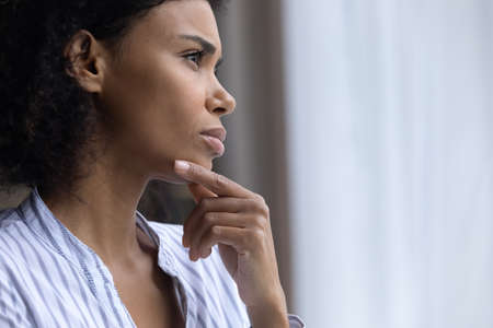 Close up of unhappy pensive young African American woman look in window distance thinking pondering. Thoughtful mixed race ethnicity female feel lonely lack communication on covid-19 lockdown at home.の写真素材