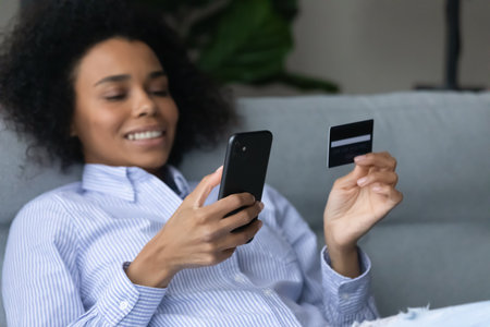 Close up of smiling African American woman relax at home shopping online on cellphone with credit debit card. Happy biracial female client buy on web on smartphone make secure internet payment.の写真素材