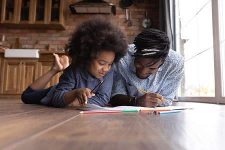 Loving happy young African American dad and teen daughter relax together draw paint in album at home. Caring ethnic father and small girl child have fun involved in art activity. Hobby concept.の写真素材