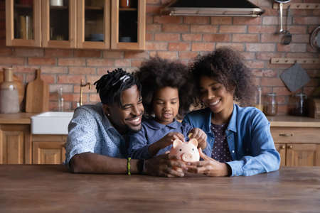 Smiling young African American family with daughter save money in piggybank feel provident economical about finances. Happy biracial parents with ethnic child make financial investment for future.の写真素材