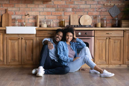 Portrait of happy ethnic couple renters tenants relax on wooden floor in renovated design kitchen enjoy weekend together. Smiling African American man and woman rest on moving day to new home.の写真素材