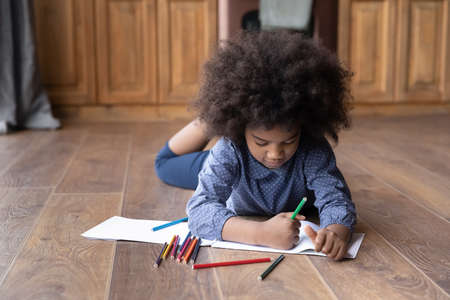 Cute small teen African American girl child lying on warm wooden kitchen floor drawing painting in...