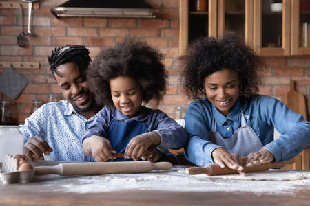 Smiling young African American family with teen daughter enjoy baking making cookies in kitchen together. Happy ethnic parents with girl child relax cooking at home, enjoy weekend. Hobby concept.の写真素材